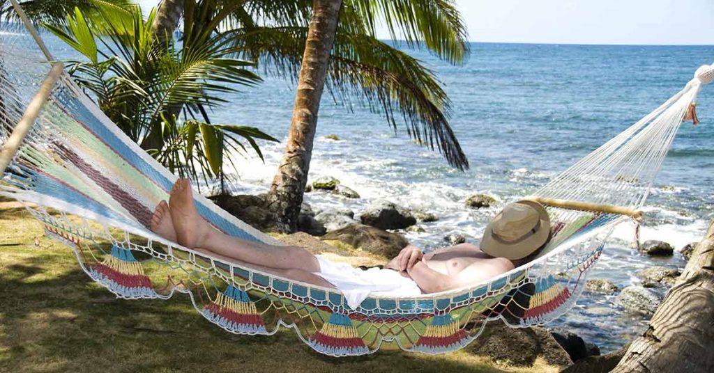 Ein Mann liegt entspannt in seiner Hängematte am Strand im Ausland unter Palmen, mit Hut auf dem Gesicht, und genießt sein Leben, weil er weiß, dass ein Qualitätsmakler sich um den Verkauf seiner Immobilie in Deutschland kümmert | Immobilienverkauf